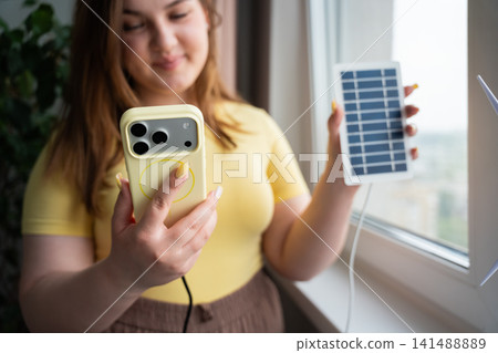 Woman using smartphone connected to portable solar charger by a window. Clean energy mobile connectivity and renewable technology Woman using smartphone connected to portable solar charger by a window. Clean energy mobile connectivity and renewable technology 141488889
