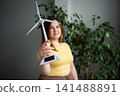 Young woman holding a small model wind turbine in a bright interior space. Sustainability education renewable energy and eco startup concepts 141488891