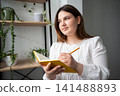 Young woman writing in a journal while sitting at home with plant-filled shelves in the background. Personal development journaling and at-home education 141488893