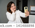 Young woman smiling while holding smartphone for a video call in a modern home setting. Social media influencer marketing and personal branding 141488898