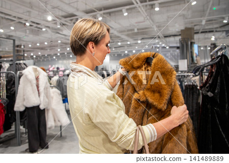 Woman choosing faux fur coat in clothing store. Customer shopping for winter apparel. Sustainable fashion and ethical clothing retail concept in boutique. Winter style selection. Woman choosing faux fur coat in clothing store. Customer shopping for winter apparel. Sustainable fashion and ethical clothing retail concept in boutique. Winter style selection. 141489889