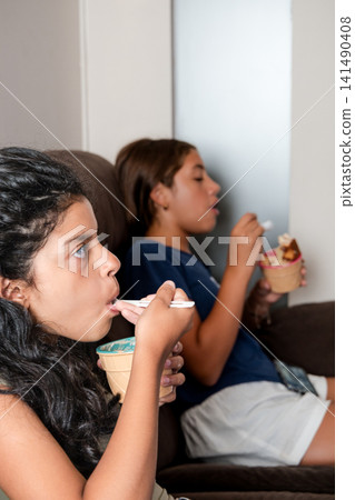 Two hispanic girls eating ice cream on sofa watching tv 141490408