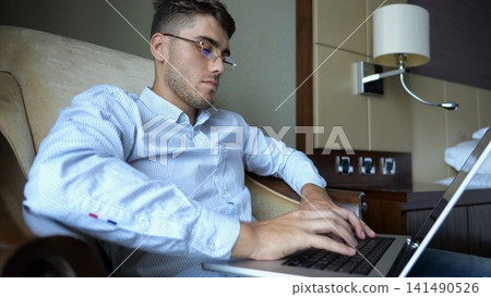 Handsome entrepreneur use notebook for work sitting in armchair at home interior. Young businessman in glasses working on laptop in modern hotel room. Manager showing remote job and productivity Handsome entrepreneur use notebook for work sitting in armchair at home interior. Young businessman in glasses working on laptop in modern hotel room. Manager showing remote job and productivity 141490526