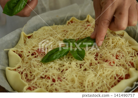 Close up of woman's hands preparing a homemade tomato tart with mustard, cream, grated cheese, tomato pieces and fresh basil leaves from the garden, showing a natural cooking process. Close up of woman's hands preparing a homemade tomato tart with mustard, cream, grated cheese, tomato pieces and fresh basil leaves from the garden, showing a natural cooking process. 141490757