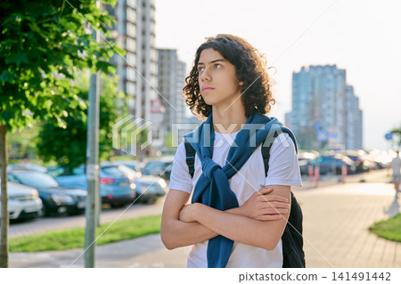 Thoughtful teenage male student with curly hair looking away on modern city street 141491442