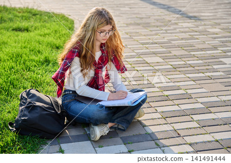 Focused young female student sitting on paving tiles and reading textbook 141491444