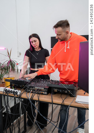 DJ in orange hoodie preparing music set at workstation with audio equipment in studio. DJ, workstation, audio equipment, music set, studio 141491836