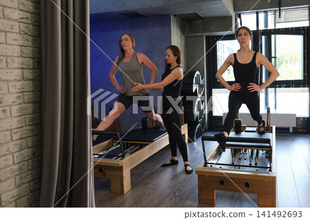 A trainer assists a woman balancing on a Pilates reformer while another client repeats the pose nearby. The scene reflects boutique fitness service, personal guidance, mobility work, and quality care. A trainer assists a woman balancing on a Pilates reformer while another client repeats the pose nearby. The scene reflects boutique fitness service, personal guidance, mobility work, and quality care. 141492693