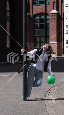 Woman playfully turning around a pole with relaxed posture and windblown hair in an urban setting. The lively scene highlights lightness, inner freedom and carefree lifestyle. Woman playfully turning around a pole with relaxed posture and windblown hair in an urban setting. The lively scene highlights lightness, inner freedom and carefree lifestyle. 141492711