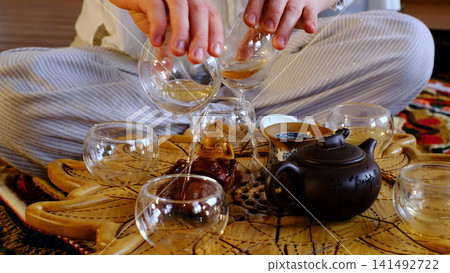 Close view of glass teacups pouring amber tea over an ornate wooden tray with a clay teapot nearby. Tea meditation and slow living create peaceful pauses for focus, balance, and relaxation. Close view of glass teacups pouring amber tea over an ornate wooden tray with a clay teapot nearby. Tea meditation and slow living create peaceful pauses for focus, balance, and relaxation. 141492722