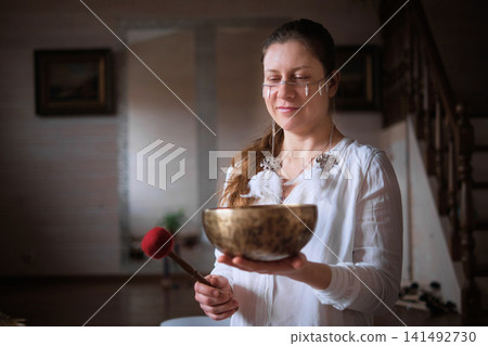 Eye level view of a serene practitioner presenting a bronze singing bowl while holding a soft mallet indoors. Vibrational sound rituals help reduce tension and restore mindful focus. Eye level view of a serene practitioner presenting a bronze singing bowl while holding a soft mallet indoors. Vibrational sound rituals help reduce tension and restore mindful focus. 141492730
