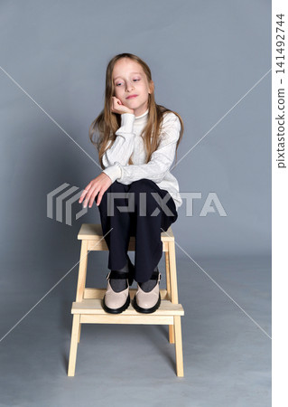 Girl sits wooden step stool with hands gently placed on her knee, subtle smile and quiet confidence Girl sits wooden step stool with hands gently placed on her knee, subtle smile and quiet confidence 141492744