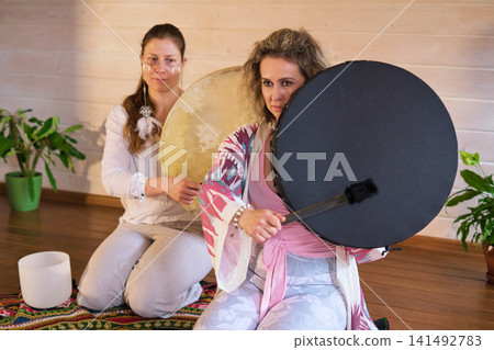 Eye level portrait of two focused women seated with large frame drums and soft mallets in a warm wooden studio. Rhythmic sound healing supports grounding, stress relief, and shared meditation. 141492783