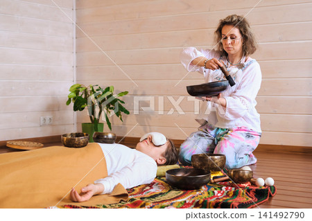 Eye level view of a private sound bath with a relaxed client lying on a colorful rug as a therapist works with singing bowls. Wellness rituals support calm focus, grounding, and deep rest. Eye level view of a private sound bath with a relaxed client lying on a colorful rug as a therapist works with singing bowls. Wellness rituals support calm focus, grounding, and deep rest. 141492790