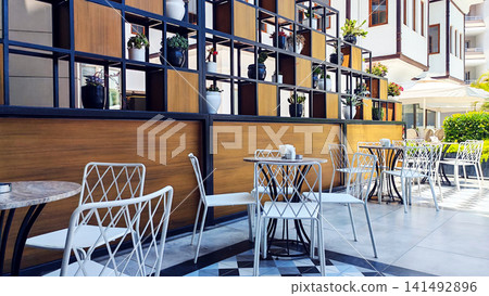 Minimalist caf terrace with empty tables and chairs in a bright hotel courtyard surrounded by plants. The peaceful travel scene reflects vacation relaxation, outdoor dining and sunny resort mornings. Minimalist caf terrace with empty tables and chairs in a bright hotel courtyard surrounded by plants. The peaceful travel scene reflects vacation relaxation, outdoor dining and sunny resort mornings. 141492896