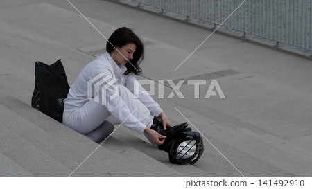 Seated on concrete steps, a woman in white secures her jump shoes. This image reflects the commitment to an active lifestyle and the preparation required for effective workouts. Seated on concrete steps, a woman in white secures her jump shoes. This image reflects the commitment to an active lifestyle and the preparation required for effective workouts. 141492910