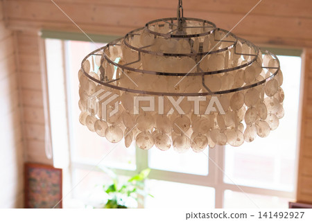 Close up of a shell chandelier hanging in front of a bright window inside a warm wooden room. Natural textures and soft daylight bring boho style, home comfort, and relaxed interior design. Close up of a shell chandelier hanging in front of a bright window inside a warm wooden room. Natural textures and soft daylight bring boho style, home comfort, and relaxed interior design. 141492927