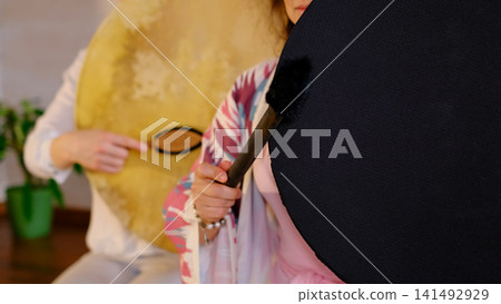 Selective focus shows a woman holding a black frame drum and padded mallet while another plays a natural skin drum behind. Rhythmic sound rituals help release tension and restore balance. Selective focus shows a woman holding a black frame drum and padded mallet while another plays a natural skin drum behind. Rhythmic sound rituals help release tension and restore balance. 141492929