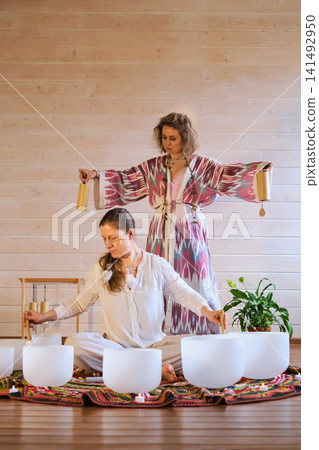 Eye level view of women using crystal bowls and wooden chimes during a candlelit sound therapy ritual on a bright rug. Mindful vibrations bring comfort, grounding, stress relief, and calm. Eye level view of women using crystal bowls and wooden chimes during a candlelit sound therapy ritual on a bright rug. Mindful vibrations bring comfort, grounding, stress relief, and calm. 141492950