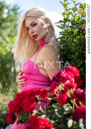 Adult blonde woman poses in profile with one hand on her waist beside vivid red roses. Elegant outdoor portrait for fashion, beauty, and floral advertising. Adult blonde woman poses in profile with one hand on her waist beside vivid red roses. Elegant outdoor portrait for fashion, beauty, and floral advertising. 141493057