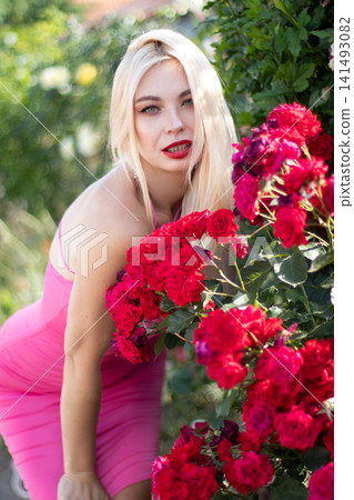 Adult blonde woman leans toward a cluster of vivid red roses and looks at the camera. Bright floral portrait for fashion, beauty, and summer lifestyle use. Adult blonde woman leans toward a cluster of vivid red roses and looks at the camera. Bright floral portrait for fashion, beauty, and summer lifestyle use. 141493082