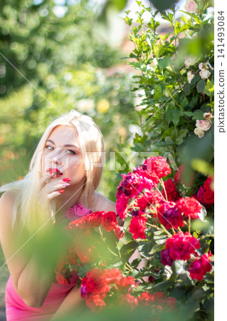 Adult blonde woman touches her lip while framed by red roses and soft foreground foliage. Expressive portrait for beauty, lipstick, and manicure themes. Adult blonde woman touches her lip while framed by red roses and soft foreground foliage. Expressive portrait for beauty, lipstick, and manicure themes. 141493084