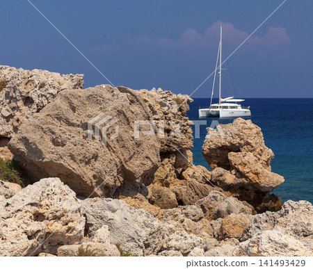 Rocky Coastline with Sailing Catamaran in the Mediterranean Sea near Rhodes, Greece Rocky Coastline with Sailing Catamaran in the Mediterranean Sea near Rhodes, Greece 141493429