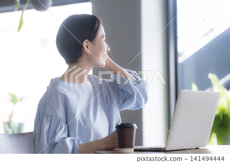 一位日本女性坐在辦公室，面前放著筆記型電腦和咖啡，目光投向窗外。這是一位職業婦女在辦公室休息時的側臉照。 141494444