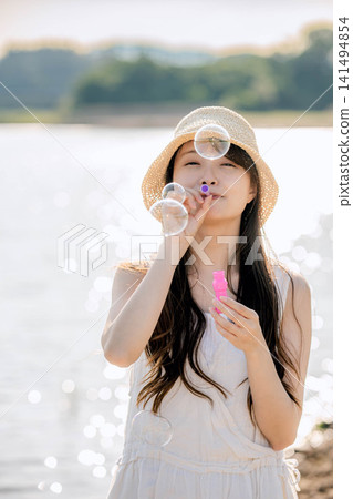 一位身穿白色洋裝的女子在湖邊快樂地吹著肥皂泡。 一位身穿白色洋裝的女子在湖邊快樂地吹著肥皂泡。 141494854