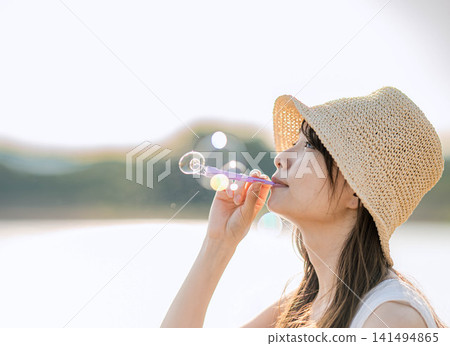 一位戴著草帽的女子在水邊吹肥皂泡——側面視角 一位戴著草帽的女子在水邊吹肥皂泡——側面視角 141494865