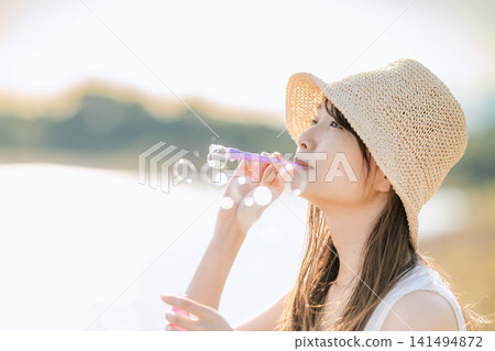 一位戴著草帽的女子在水邊吹肥皂泡。 141494872