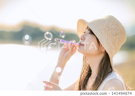 一位身穿白色洋裝的女子在湖邊逆光吹肥皂泡。 一位身穿白色洋裝的女子在湖邊逆光吹肥皂泡。 141494874