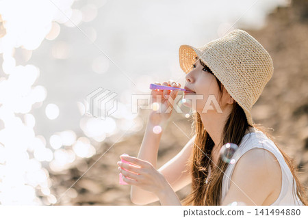一位戴著草帽的女子在夏日的陽光下吹起肥皂泡。 一位戴著草帽的女子在夏日的陽光下吹起肥皂泡。 141494880
