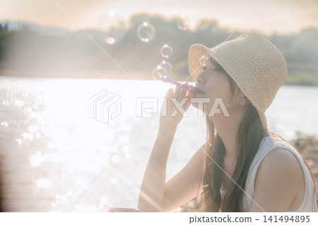 一位身穿白色洋裝的女子對著背光吹肥皂泡。 一位身穿白色洋裝的女子對著背光吹肥皂泡。 141494895