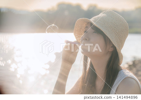一位戴著草帽的女子在夢幻般的燈光下吹著肥皂泡。 一位戴著草帽的女子在夢幻般的燈光下吹著肥皂泡。 141494896