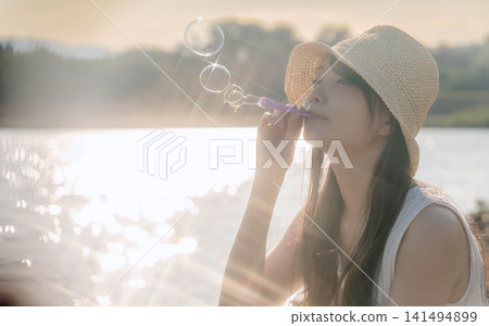 一位戴著草帽的女子正在吹肥皂泡,肥皂泡在逆光下閃閃發光。 一位戴著草帽的女子正在吹肥皂泡,肥皂泡在逆光下閃閃發光。 141494899