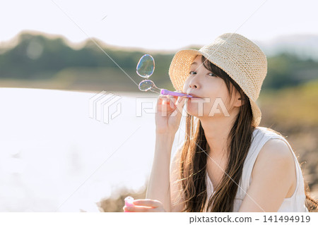 一位戴著草帽的女子在綠意盎然的背景下吹著肥皂泡。 一位戴著草帽的女子在綠意盎然的背景下吹著肥皂泡。 141494919
