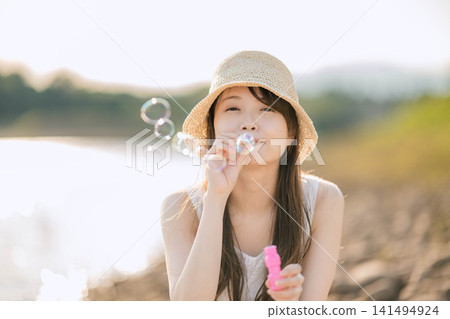 一位女子在陽光普照的湖邊吹肥皂泡。 一位女子在陽光普照的湖邊吹肥皂泡。 141494924
