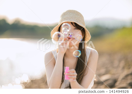 一位戴著草帽的女子在夏日陽光下吹起肥皂泡。 一位戴著草帽的女子在夏日陽光下吹起肥皂泡。 141494926