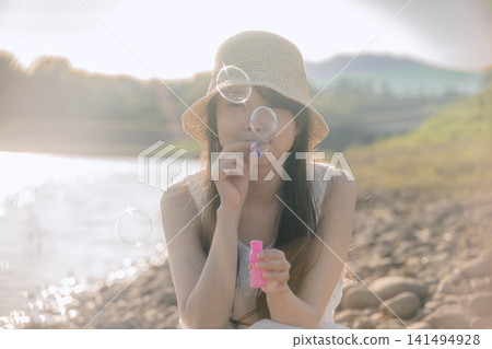 一位身穿白色洋裝的女子對著背光吹肥皂泡。 一位身穿白色洋裝的女子對著背光吹肥皂泡。 141494928