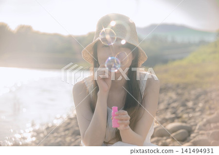 一位戴著草帽的女子在閃爍的陽光下吹著肥皂泡。 一位戴著草帽的女子在閃爍的陽光下吹著肥皂泡。 141494931