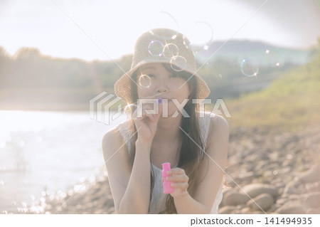 一位戴著草帽的女子正在吹肥皂泡,肥皂泡在逆光下閃閃發光。 一位戴著草帽的女子正在吹肥皂泡,肥皂泡在逆光下閃閃發光。 141494935