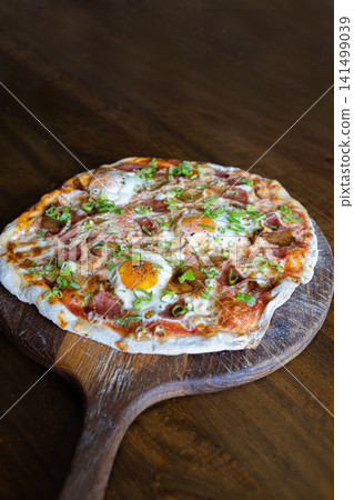 Fresh artisan breakfast pizza topped with sunny side eggs, mushrooms, cured meat, mozzarella cheese and green onions served on a rustic wooden pizza peel. Fresh artisan breakfast pizza topped with sunny side eggs, mushrooms, cured meat, mozzarella cheese and green onions served on a rustic wooden pizza peel. 141499039