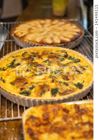 Fresh spinach mushroom quiche with apple tart and baked lasagna displayed on cooling racks in a rustic artisan bakery. Fresh spinach mushroom quiche with apple tart and baked lasagna displayed on cooling racks in a rustic artisan bakery. 141499115