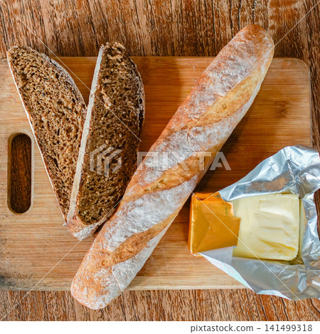 Top view of artisan baguette, sliced whole grain bread, and butter on a rustic wooden cutting board. Authentic bakery scene featuring healthy bread, breakfast, homemade baking, and natural food style 141499318