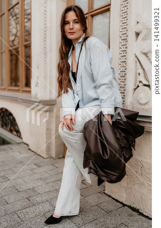 Woman urban fashion lady in blue shirt and jeans poses against classical building holding leather jacket in the city Woman urban fashion lady in blue shirt and jeans poses against classical building holding leather jacket in the city 141499321