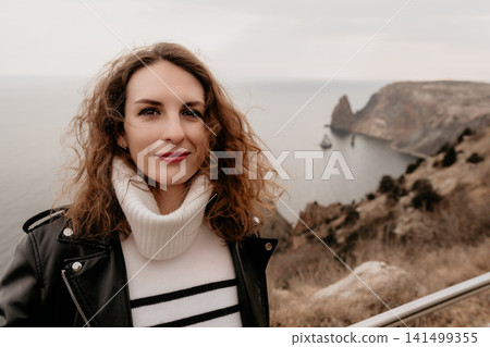 Woman leather portrait smiling on a rocky cliff overlooking the sea during an overcast day in autumn Woman leather portrait smiling on a rocky cliff overlooking the sea during an overcast day in autumn 141499355