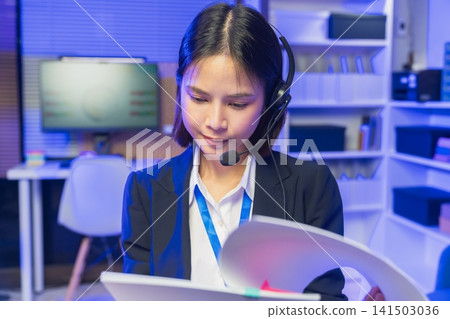 Smiling young Asian customer service representative wearing microphone headset and working on laptop Smiling young Asian customer service representative wearing microphone headset and working on laptop 141503036