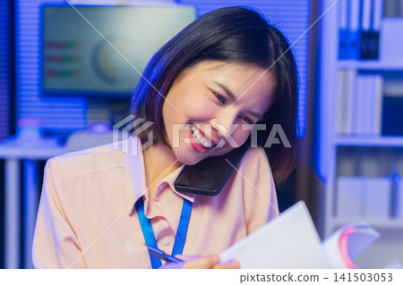 Young Asian businesswoman talking on smartphone while reviewing documents in office. Young Asian businesswoman talking on smartphone while reviewing documents in office. 141503053