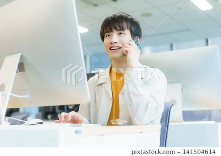 A young Japanese man is typing on his desktop computer while talking on his smartphone. A young Japanese man is typing on his desktop computer while talking on his smartphone. 141504614